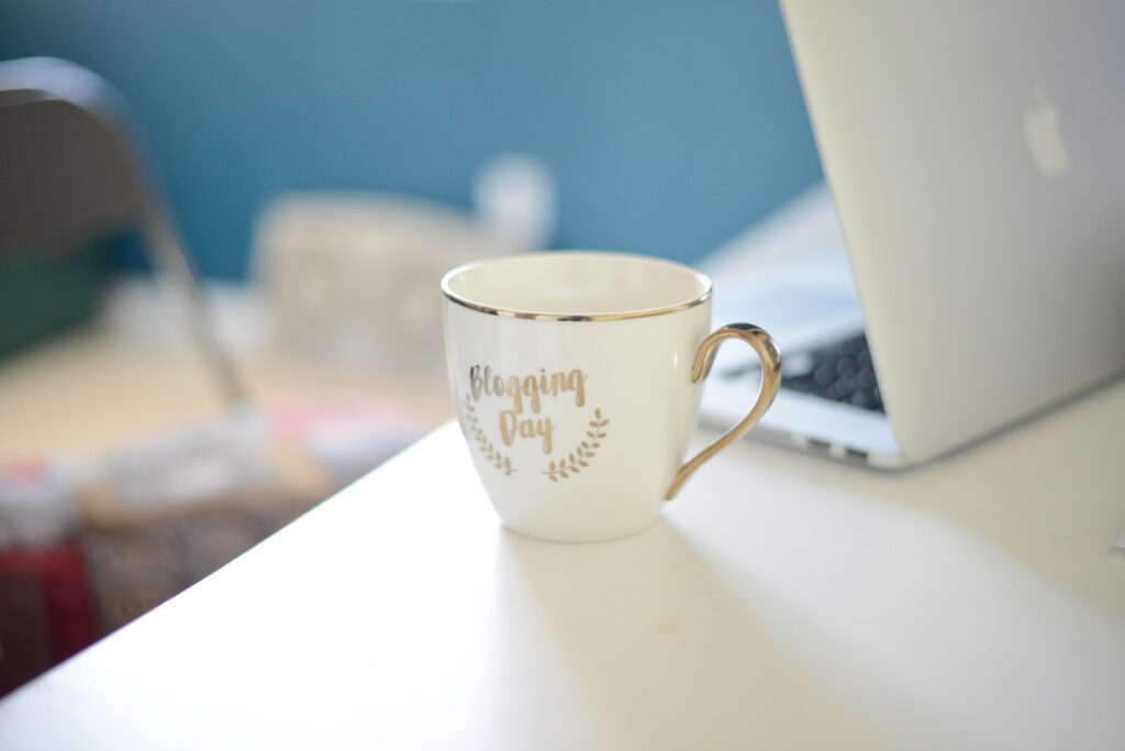 Close-up of a ceramic coffee cup labeled 'Blogging Day' on a white desk beside a laptop at a home office. Blogging