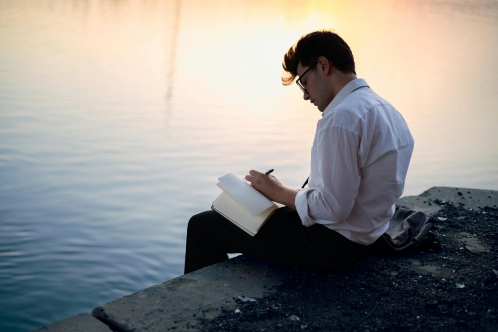 Man in white shirt writing in a notebook by the water at sunset, creating a serene and contemplative scene.