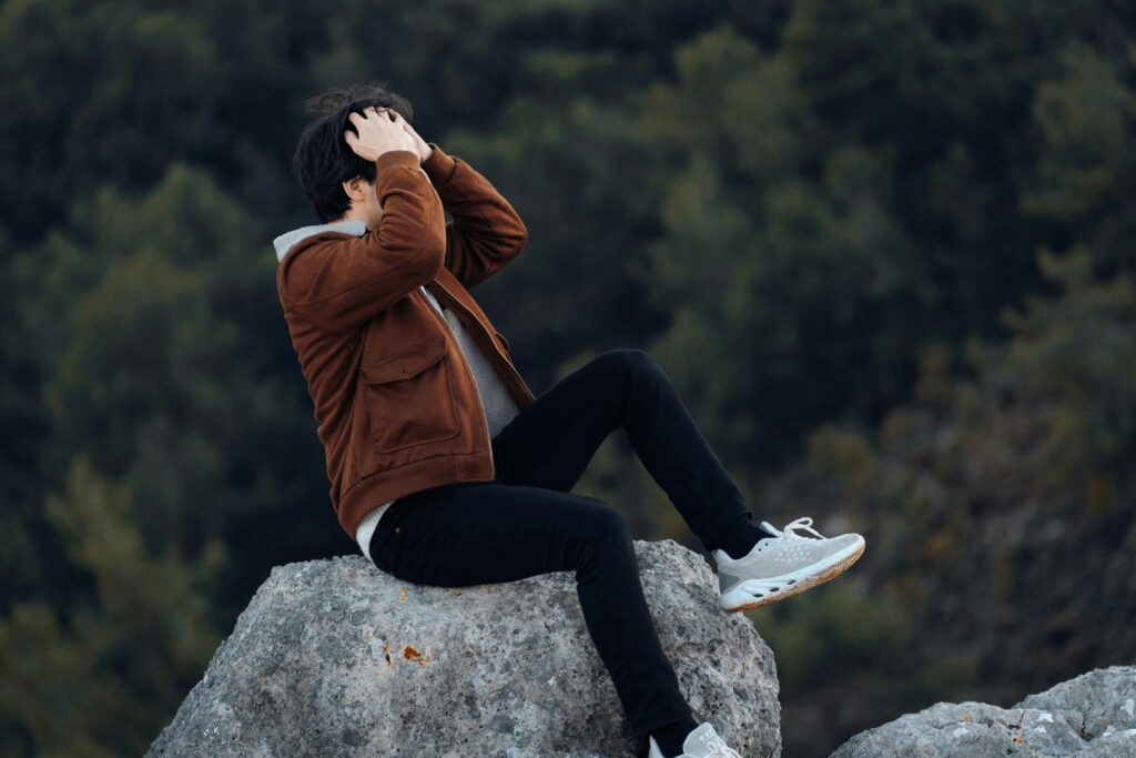 A man sits on a rock in a natural landscape, hands on head.