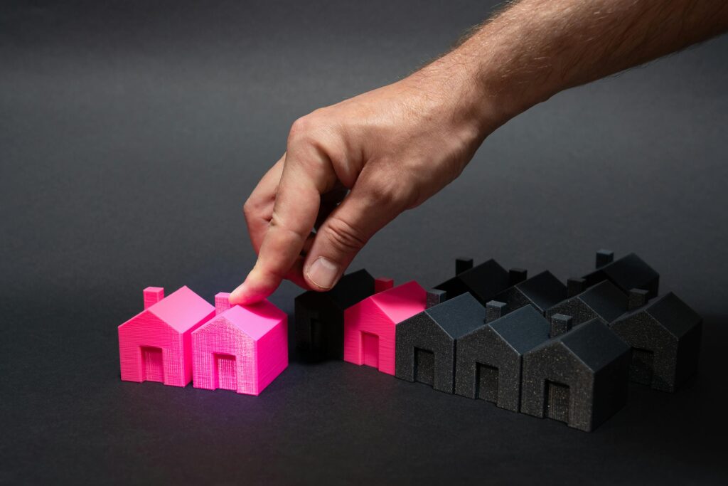A hand choosing a vibrant pink house model among black miniatures, symbolizing real estate selection. Income Sources Diversification