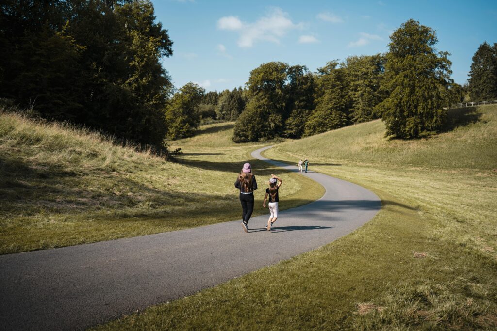 Two people walking on a scenic pathway in Woodstock, UK, surrounded by lush greenery on a bright summer day.