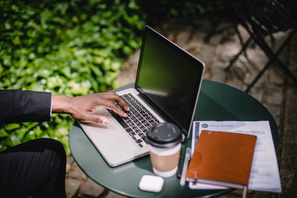 From above of crop faceless black businessman typing on laptop keyboard while working remotely at round table with takeaway coffee and notepad in outdoor cafe