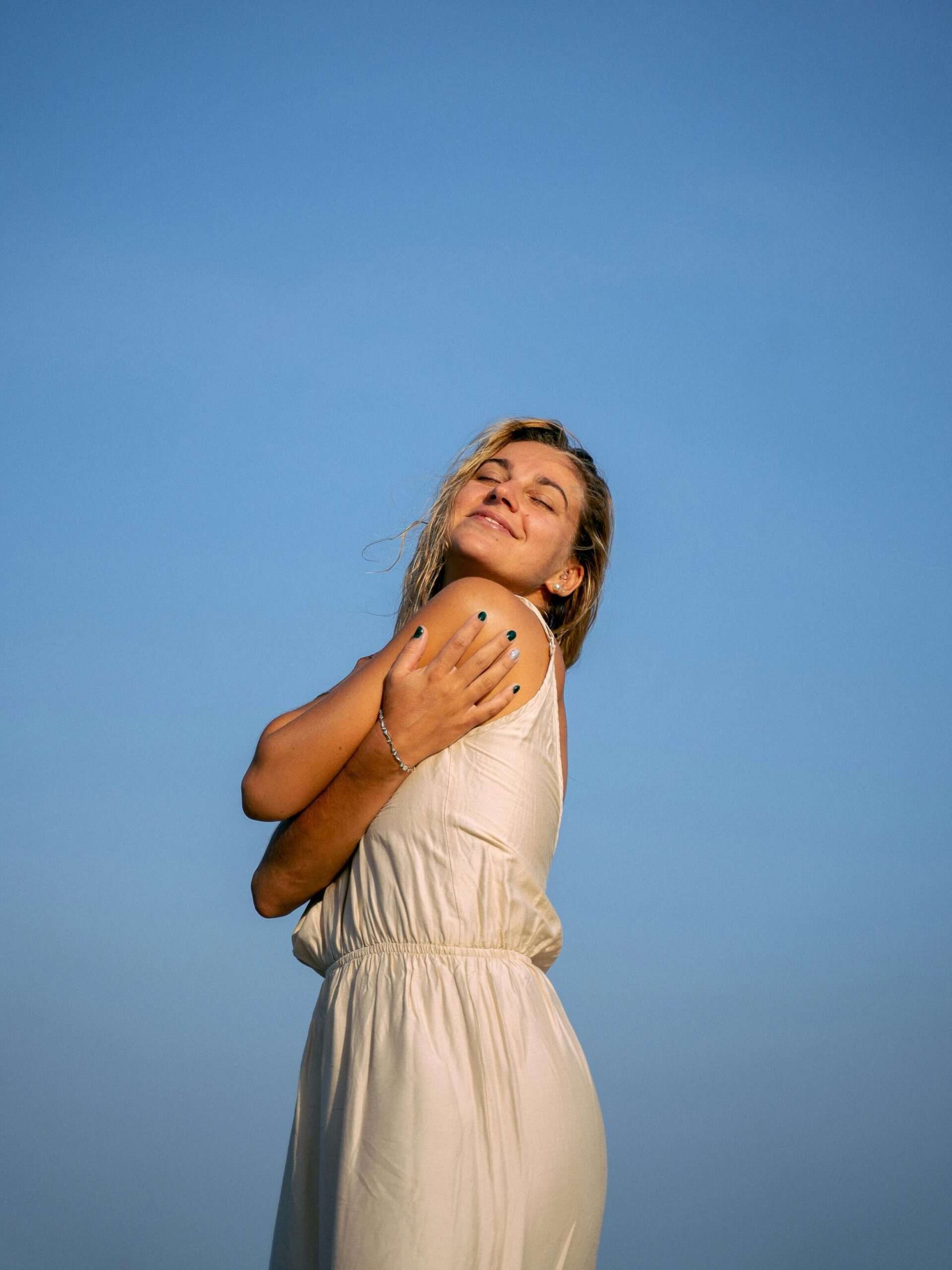 Woman in white dress embracing sunlight with closed eyes against blue sky.