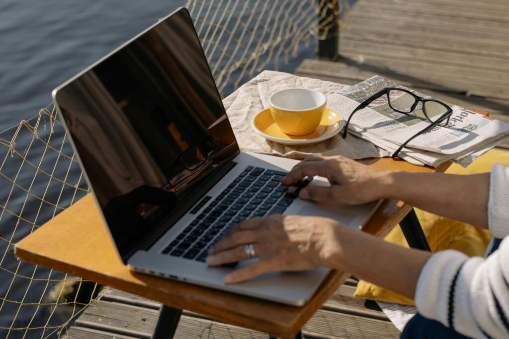 A person typing on a laptop outdoors by a lakeside with coffee and newspaper, ideal for remote work imagery. Freelancing