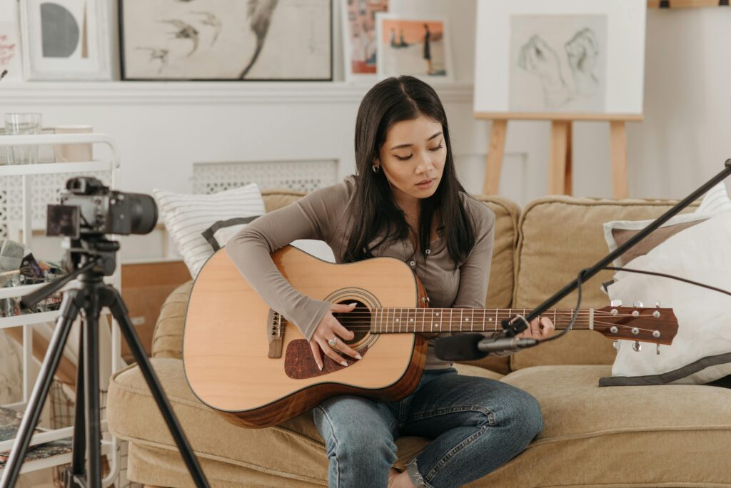 Asian woman strumming acoustic guitar indoors with camera setup. Calm and creative atmosphere.