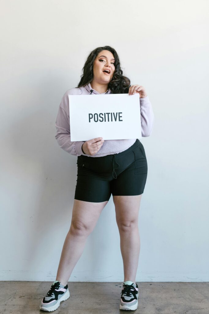 Portrait of a smiling woman posing with a sign reading 'Positive' on a white background.