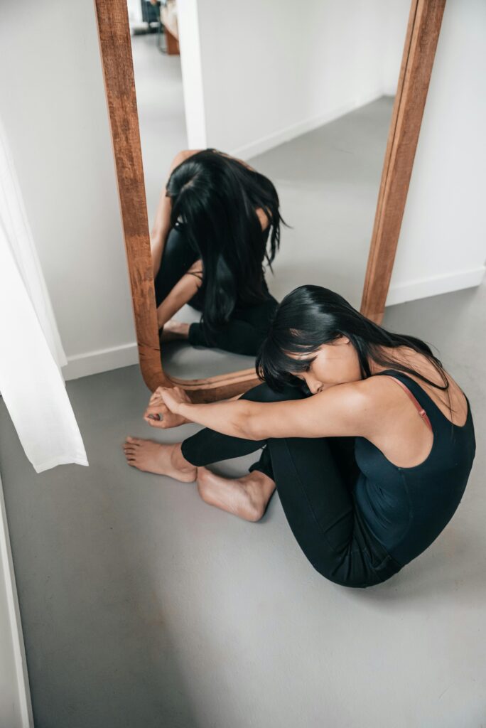 A woman sits thoughtfully by a mirror, barefoot and introspective indoors.