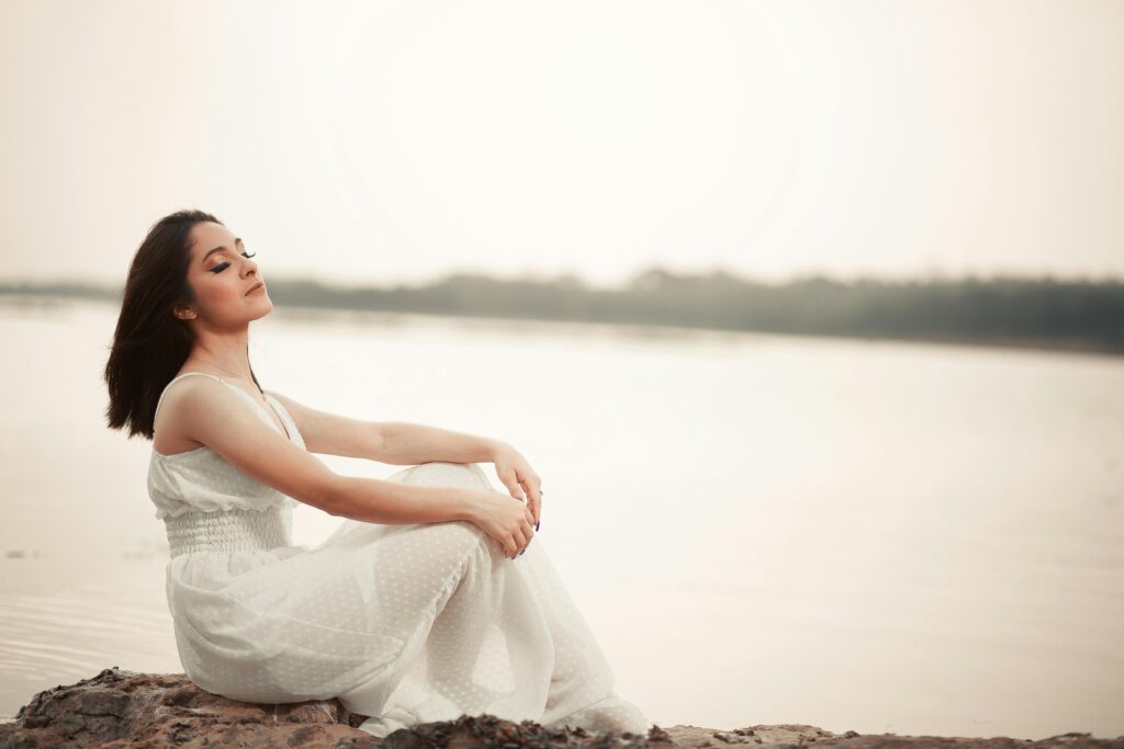 Woman in white dress sitting by water, eyes closed, enjoying tranquility.