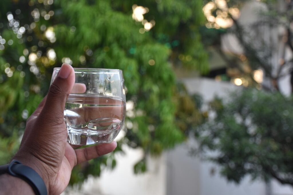 A hand holds a glass of water against a blurred green leafy background.