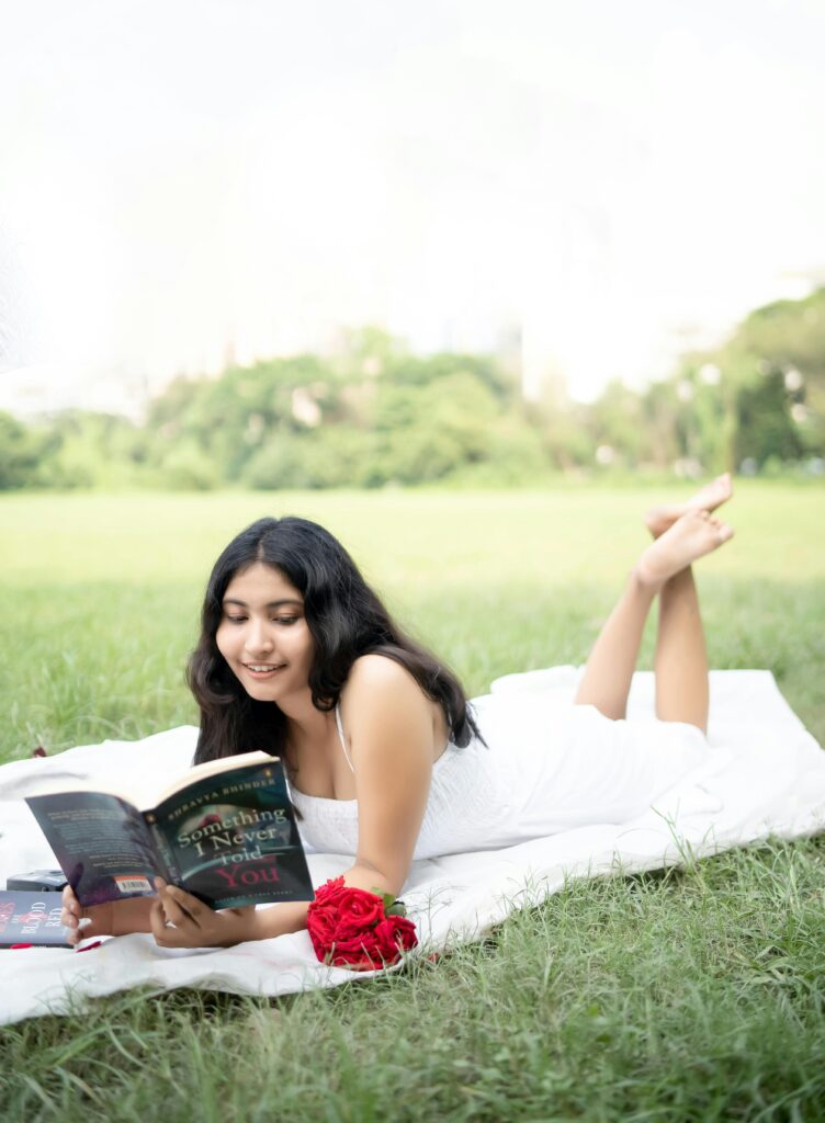 Young woman in a white dress reading a book on a blanket in a sunny park setting.