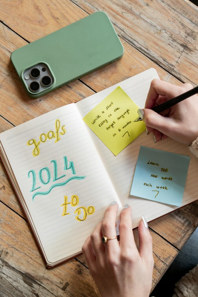 A woman writes goals in a notebook with a smartphone and sticky notes nearby.