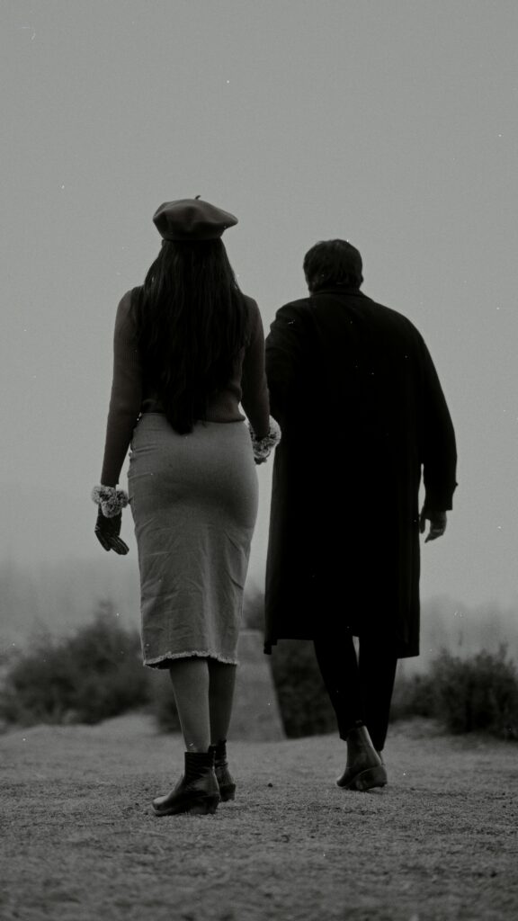 A couple holding hands walks on a beach in a monochrome setting, capturing a romantic silhouette.