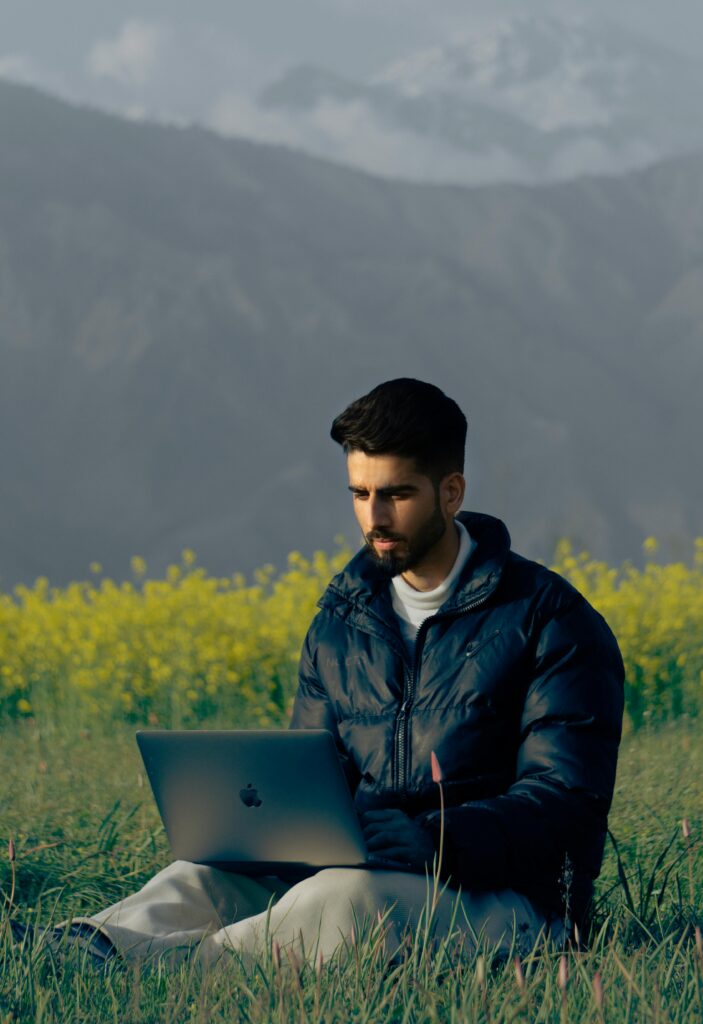 A man sitting in a picturesque field using a laptop with mountains in the background. Ideal for remote work concepts.
Apni Value Kaise Badhaye
