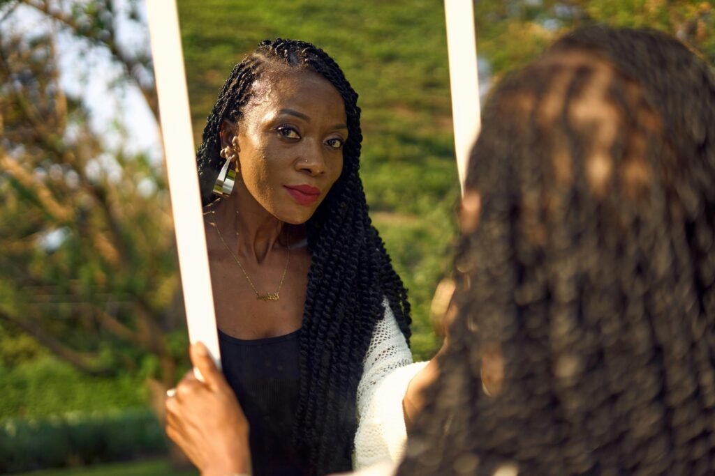 A beautiful African woman with braided hair looks in a mirror outdoors, with sunlight enhancing her natural beauty.