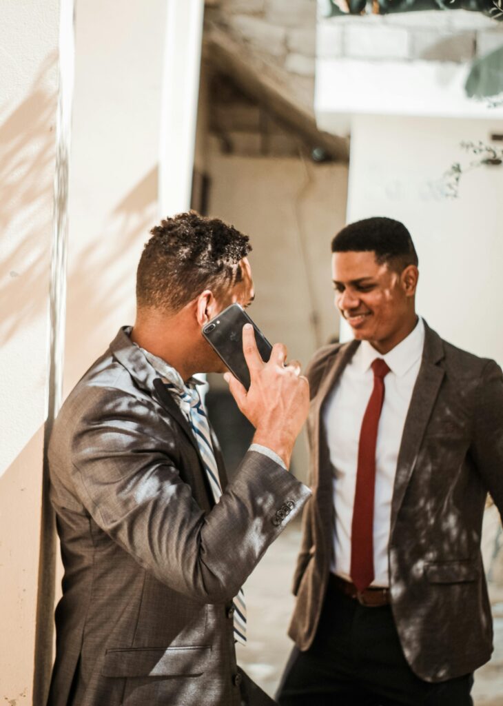 Two businessmen in corporate suits having a conversation outdoors, one smiling and the other on a phone call.