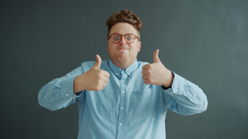 Smiling adult in glasses and blue shirt showing thumbs up indoors.