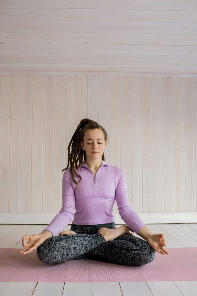 A woman sits cross-legged on a pink yoga mat meditating indoors. Calm and zen atmosphere.