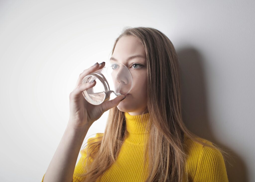 Young woman in a yellow top, drinking water from a glass against a white wall.