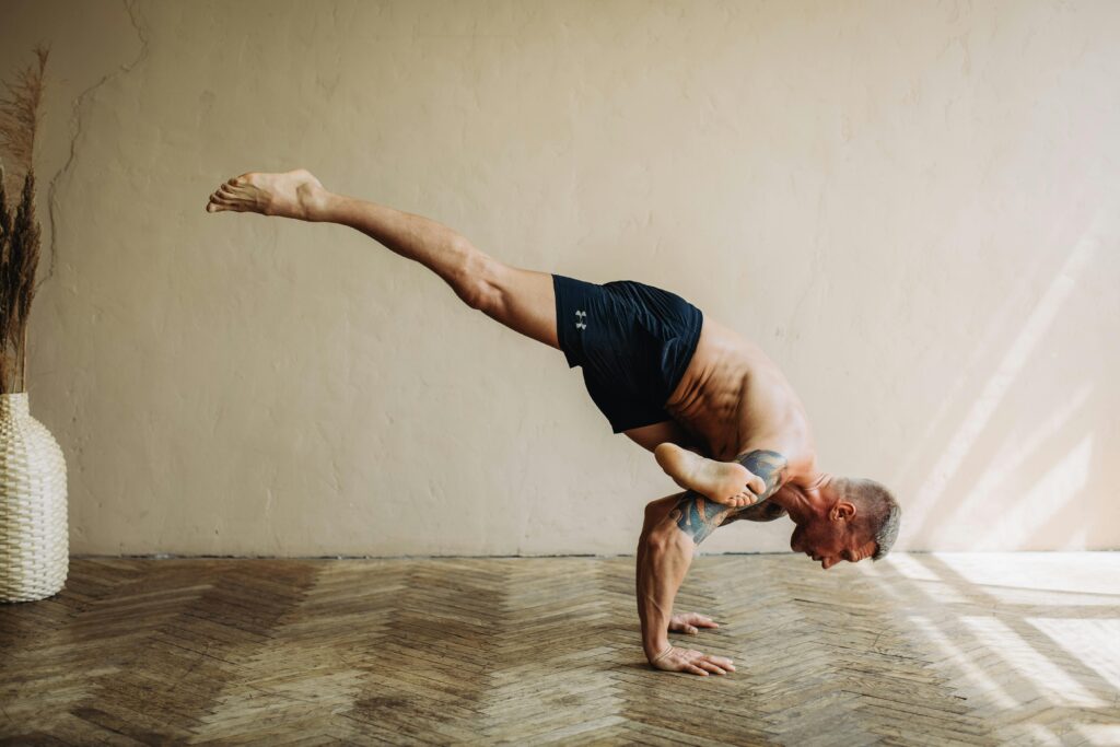 A shirtless man demonstrates advanced yoga balance indoors, showcasing strength and flexibility in a handstand position.