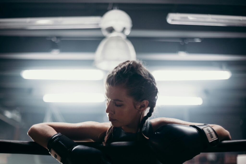 A female boxer wearing gloves rests on the ropes during an indoor training session.