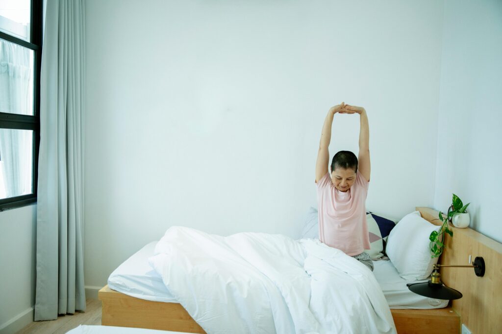 Adult Asian woman with dark short hair in casual clothes stretching arms while sitting on bed after awakening at home
