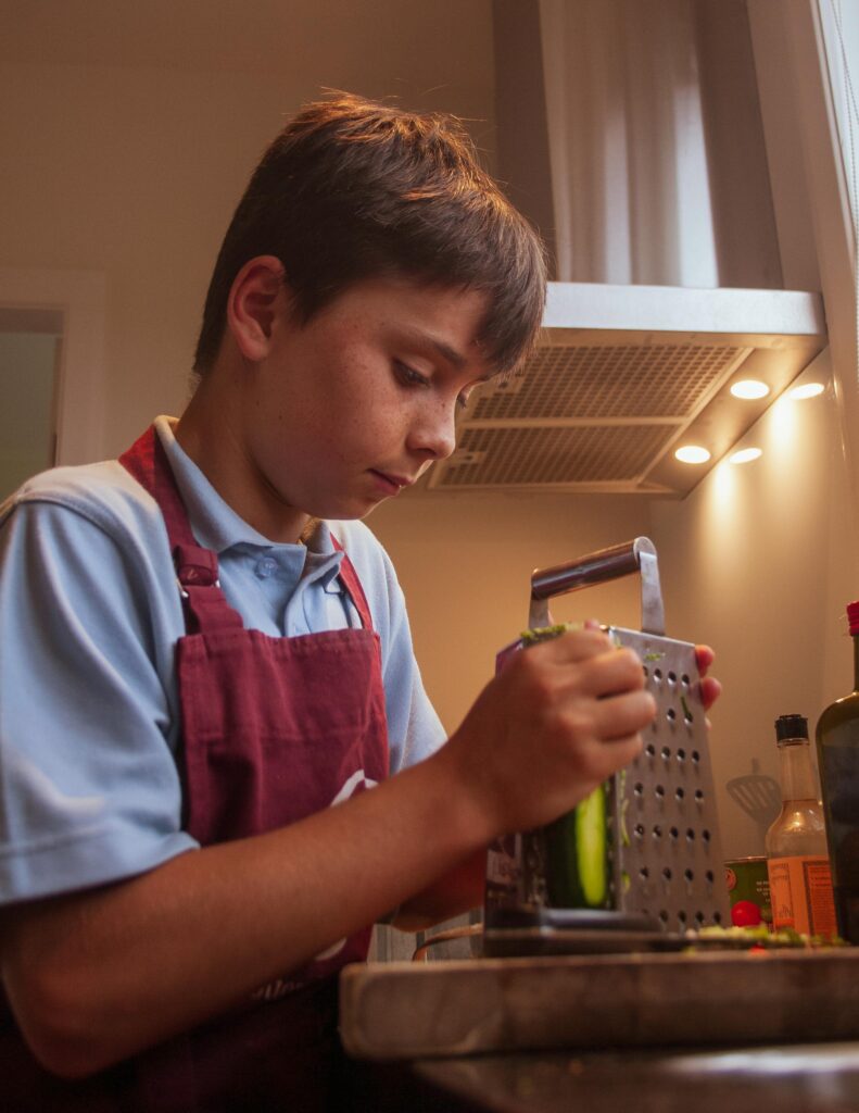 A young boy uses a grater to prepare vegetables in a well-lit kitchen environment.