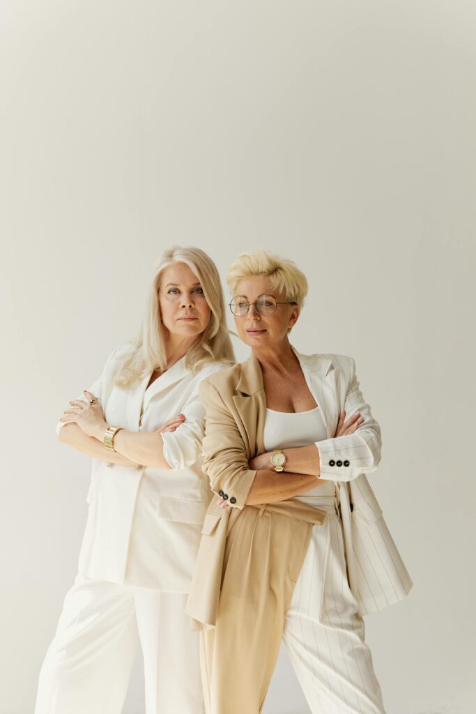 Two adult businesswomen in stylish beige and white attire confidently posing indoors.