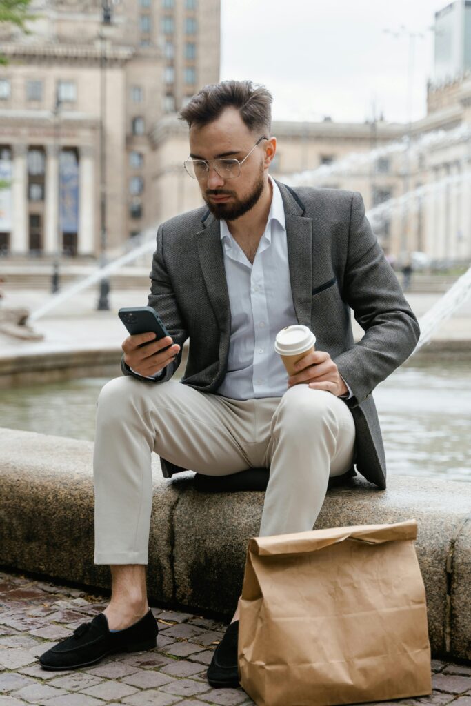 Man in a suit sitting by a city fountain with smartphone and coffee