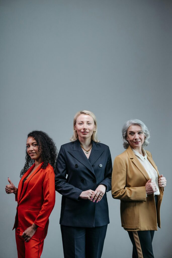 Three diverse professional women confidently posing in elegant business suits with a neutral gray background in a studio setting.