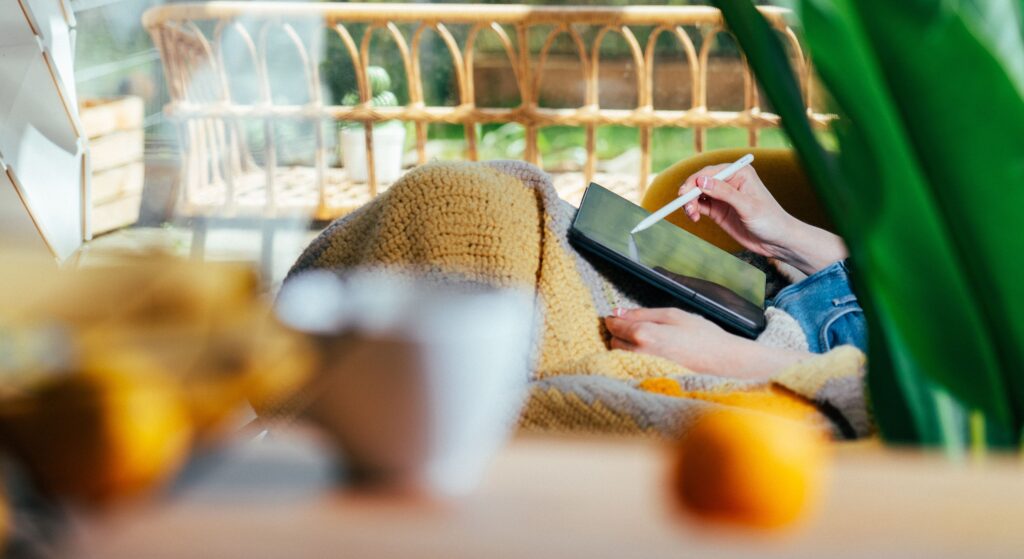 A person uses a digital pen on a tablet while wrapped in a cozy blanket indoors.