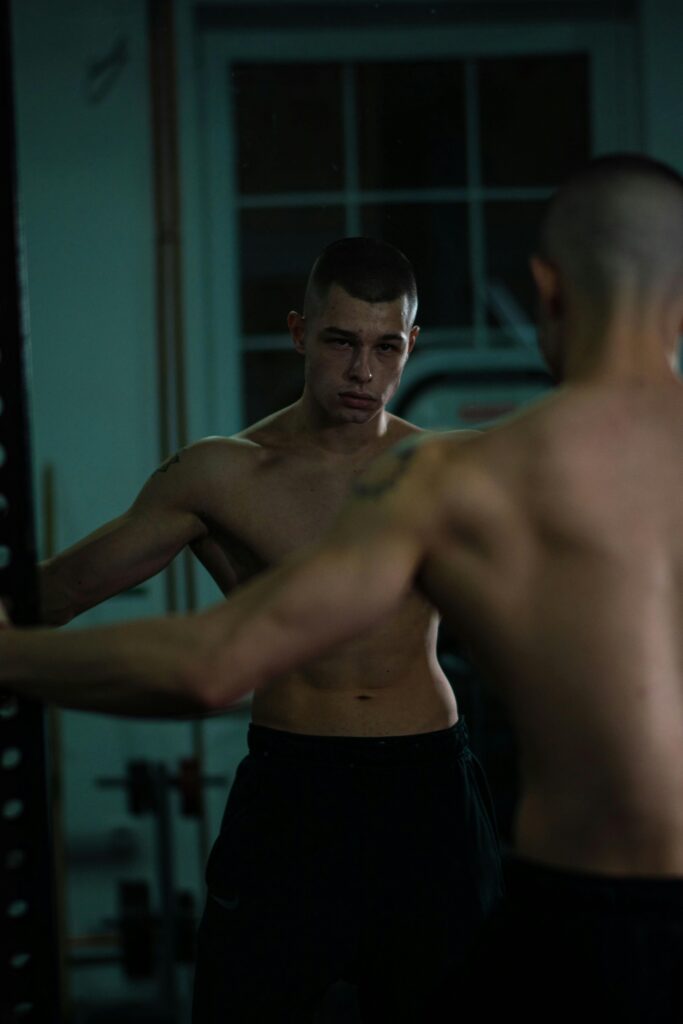 Muscular man with short hair stands topless, reflecting seriously in a gym mirror.