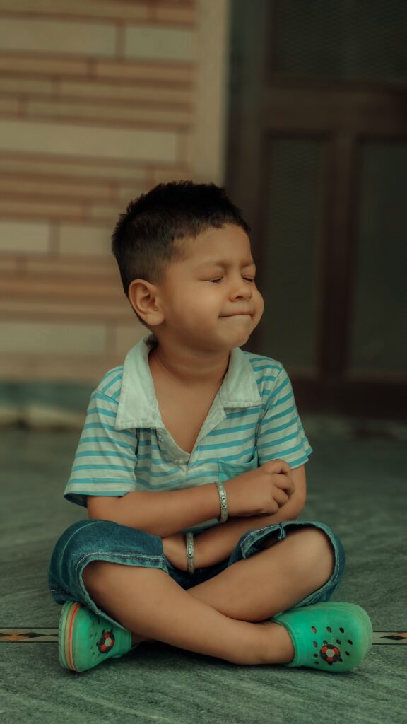 A young South Asian boy meditates with a calm expression while sitting indoors.