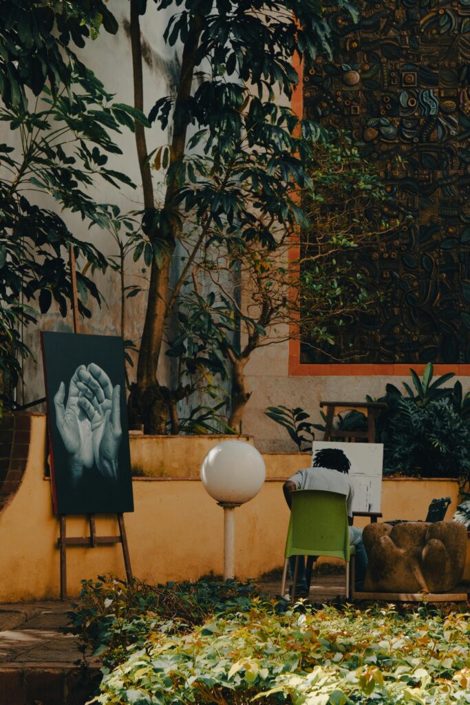 An artist paints outdoors in a lush garden with nearby artwork on display.