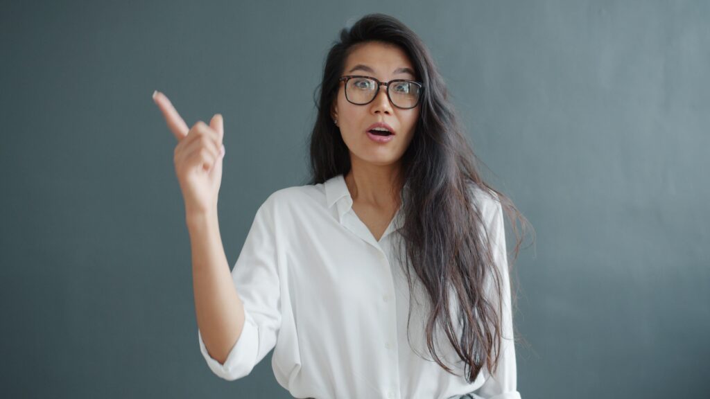 A surprised woman with glasses points upwards against a neutral background, wearing a white blouse.
