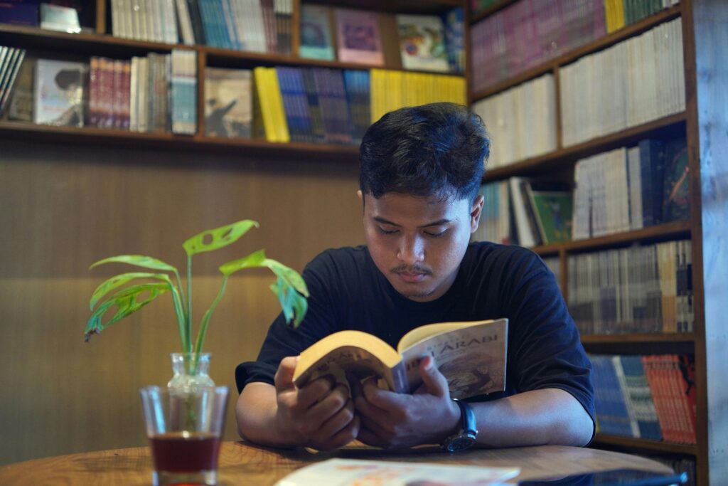 A young man engrossed in reading a book in a quiet library with bookshelves filled with literature.