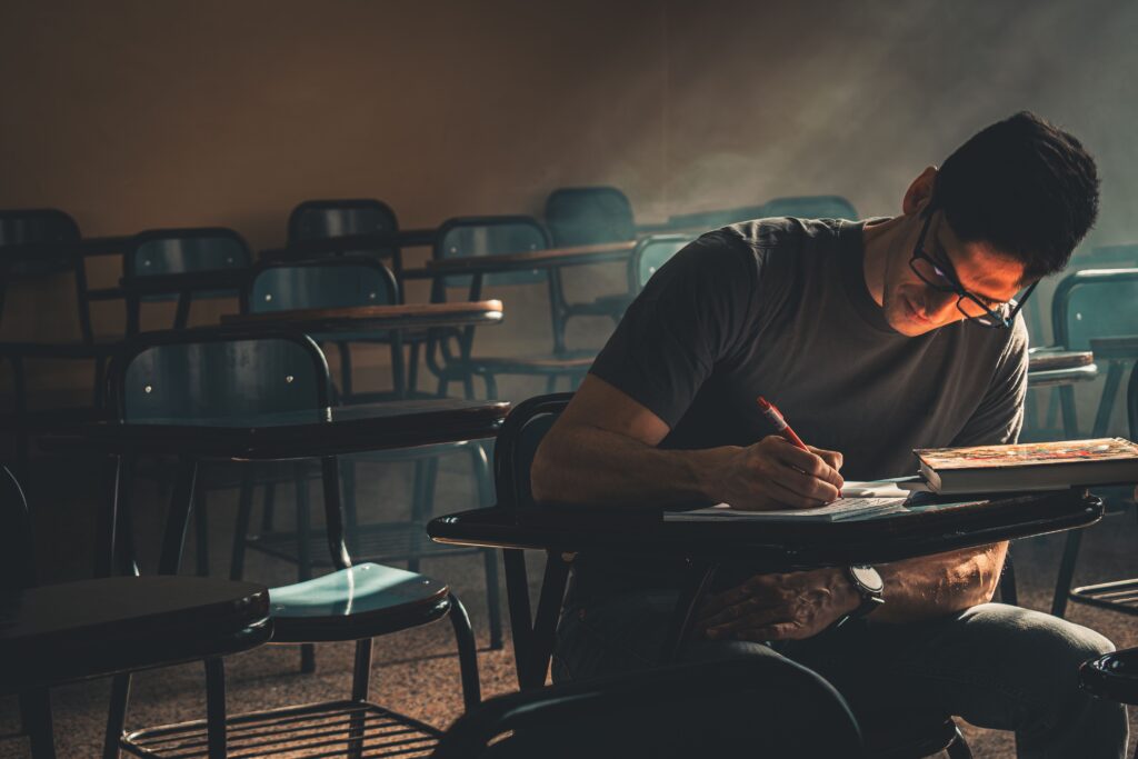 Student focused on writing, studying in a dimly lit empty classroom.
