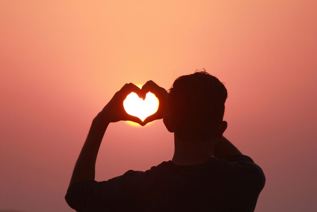 Back view silhouette of unrecognizable man in casual clothing looking at sunset and showing hands in shape of heart against sun