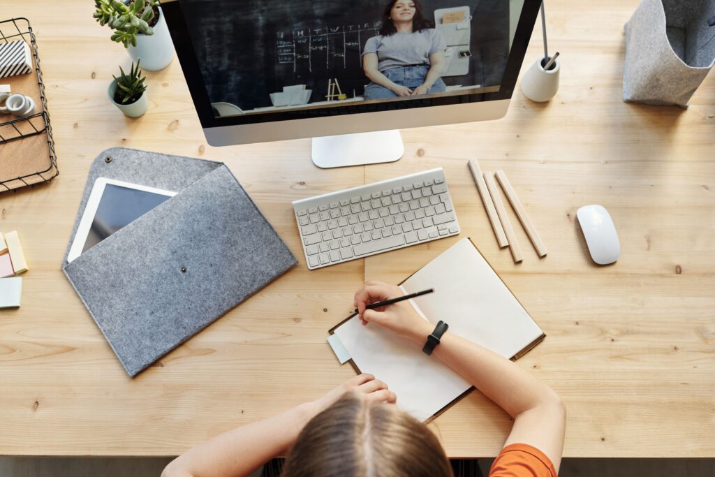 A teenager studying from home using a computer and notebook in a well-organized workspace.