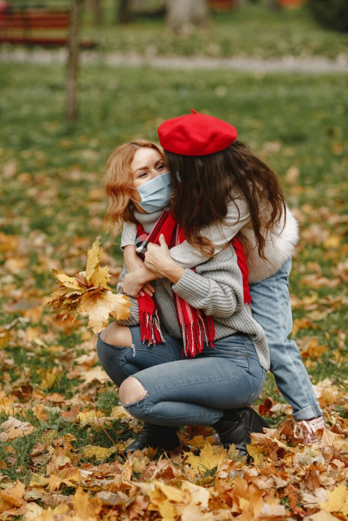 A mother and daughter wearing masks embrace in a park filled with autumn leaves, conveying warmth and protection.