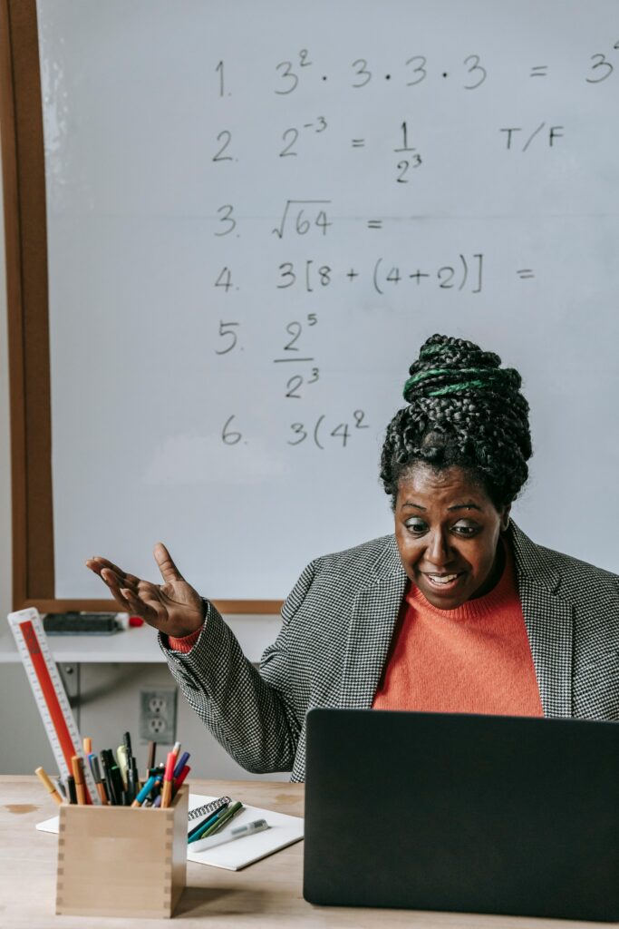 Positive smart African American female tutoring online with netbook in classroom with whiteboard