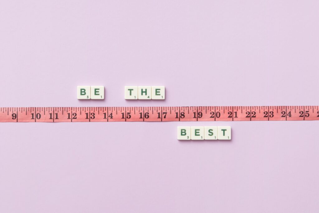 Scrabble tiles spell 'Be the Best' next to a measuring tape on a pastel background.Average Se Exceptional Kaise Bane