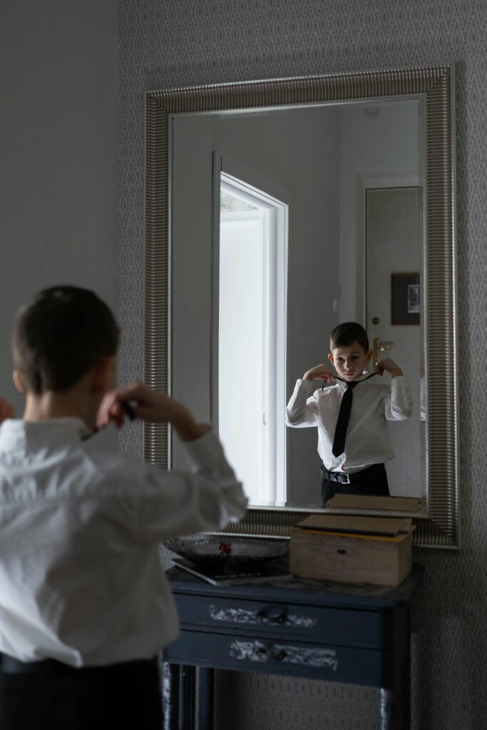 A young boy adjusting his necktie in front of a mirror, preparing for a formal occasion.
