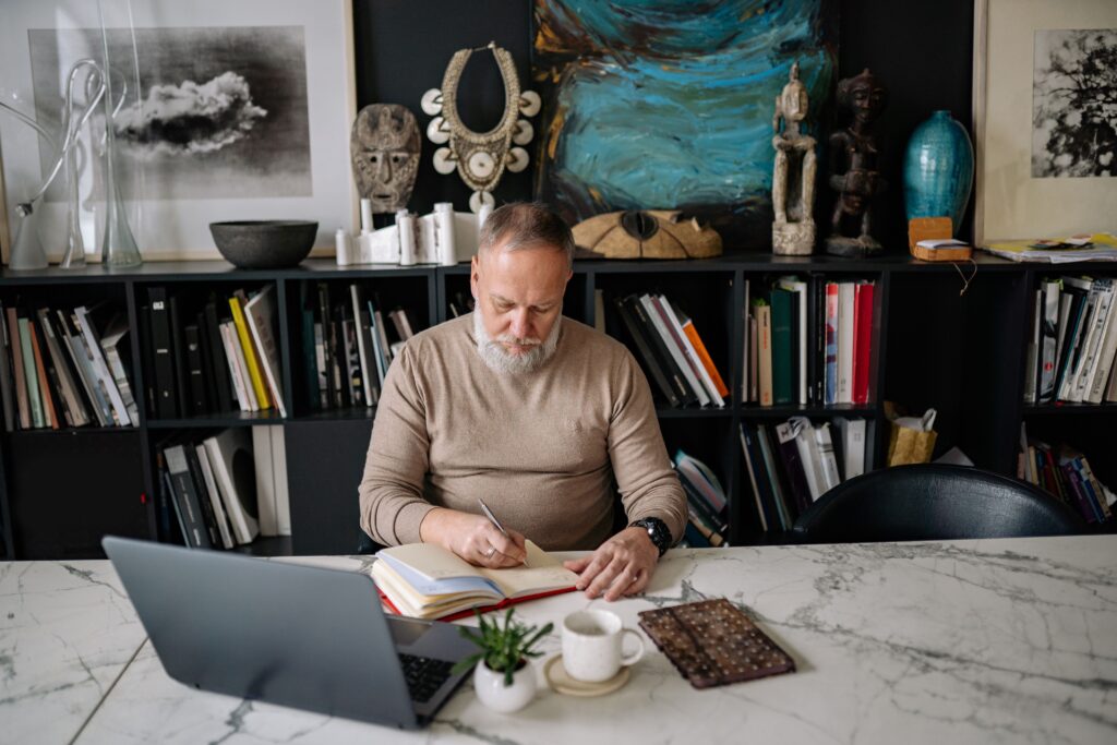 An elderly man with a beard writing in a notebook at a home office desk, surrounded by art and books.
