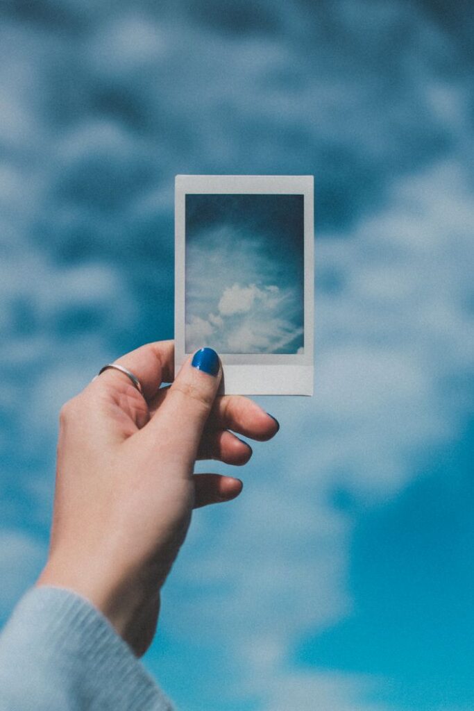 A hand with painted nails holds a Polaroid picture of clouds against a vibrant blue sky background.
Instant Gratification Se Kaise Bache?