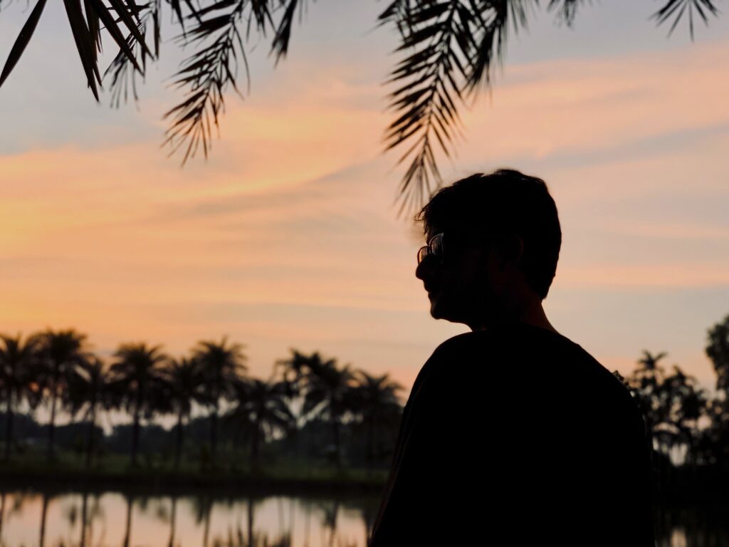 A serene sunset view with a silhouette of a man by a palm-lined lake, offering a calm and contemplative scenery.