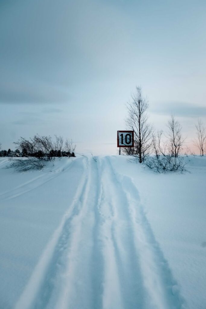 Perspective view of narrow wheels traces on snowy frozen valley with leafless trees under blue sky in winter countryside