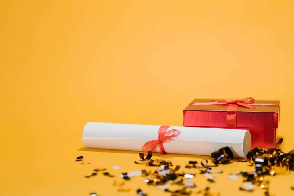 A red gift box and diploma with ribbons on a vibrant yellow background symbolizing celebration and achievement.
