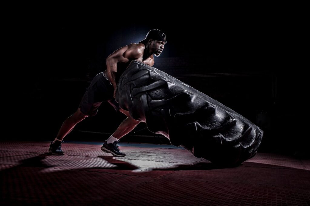 Side view full length muscular determined African American sportsman with naked torso flipping heavy truck tire in dark modern studio